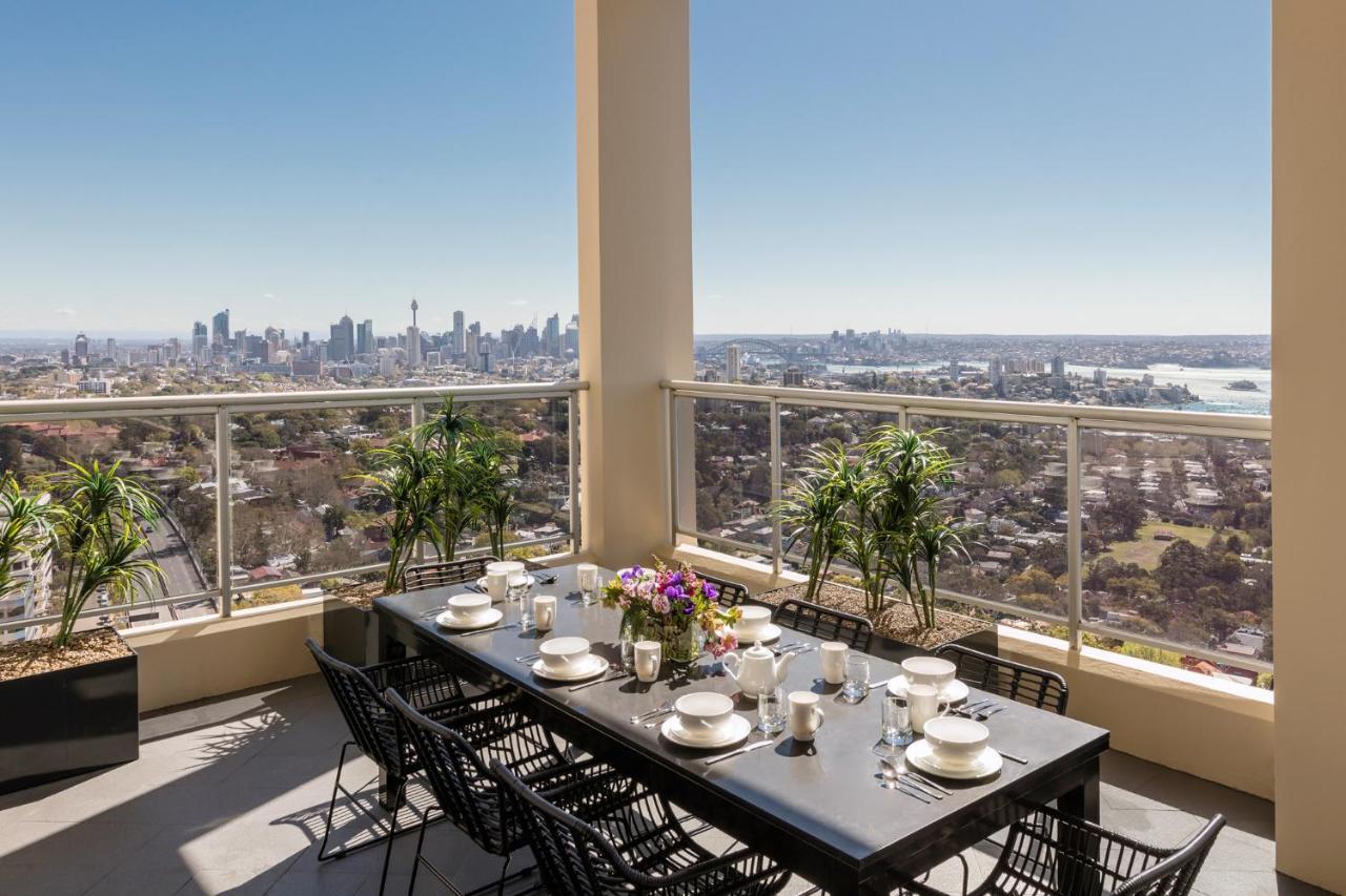 Meriton Suites Bondi Junction Sidney Exterior foto The photo shows a beautifully arranged outdoor dining area on a balcony. There is a long black dining table set for a meal, with plates, glasses, and decorative items like a vase of flowers. Surrounding the table are stylish black chairs. In the back