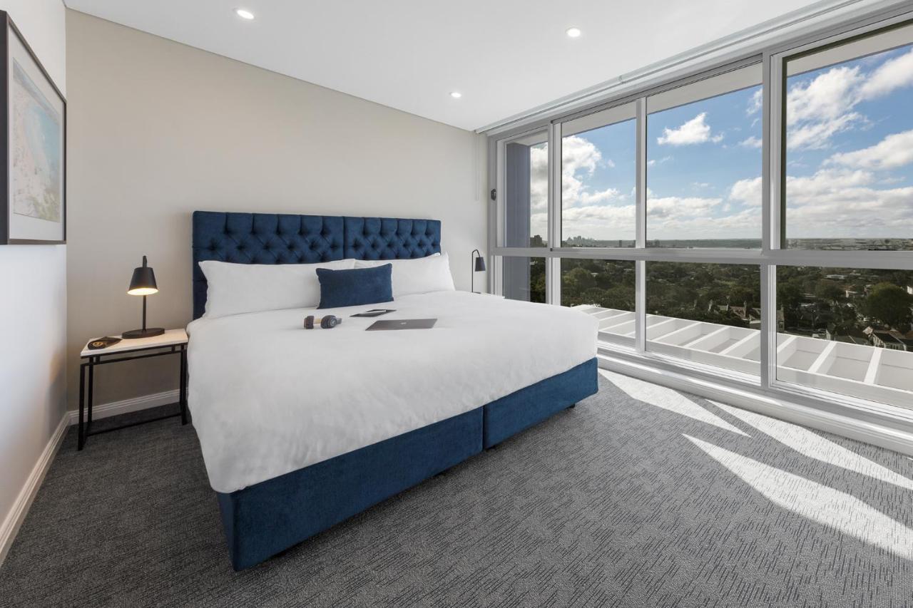 Meriton Suites Bondi Junction Sidney Exterior foto The photo shows a modern and stylish hotel room. It features a comfortable queen-size bed with a blue tufted headboard. The bed is neatly made with white linens and pillows. Next to the bed, there is a small bedside table with a desk lamp. 

On the f