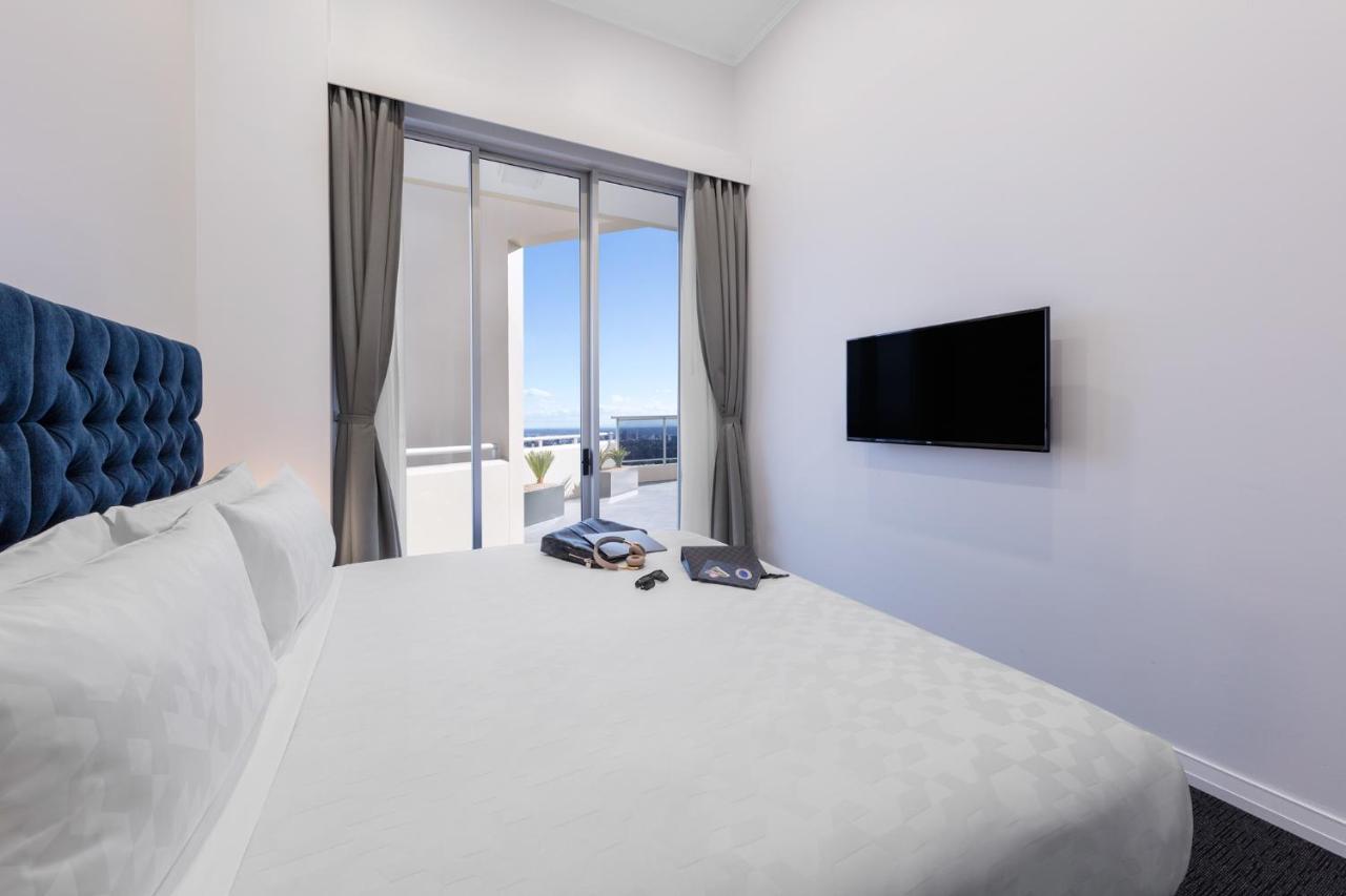 Meriton Suites Bondi Junction Sidney Exterior foto The photo shows a modern hotel room with a large, comfortable bed featuring a blue tufted headboard. There are light-colored walls, and the room is decorated simply. A flat-screen TV is mounted on the wall opposite the bed. The room has large glass d