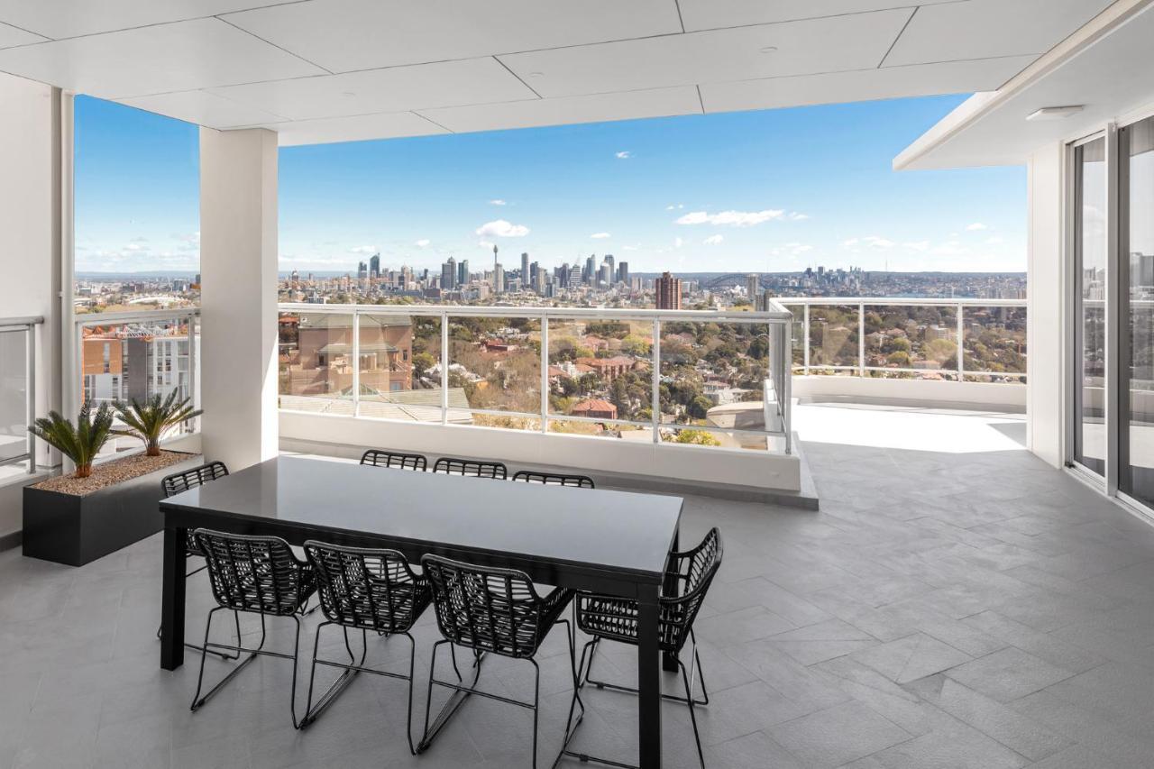 Meriton Suites Bondi Junction Sidney Exterior foto The photo shows a modern outdoor balcony or terrace space with a dining table and chairs. The table is black and has a minimalist design, surrounded by stylish chairs. In the background, there is a stunning view of a city skyline under a clear blue s